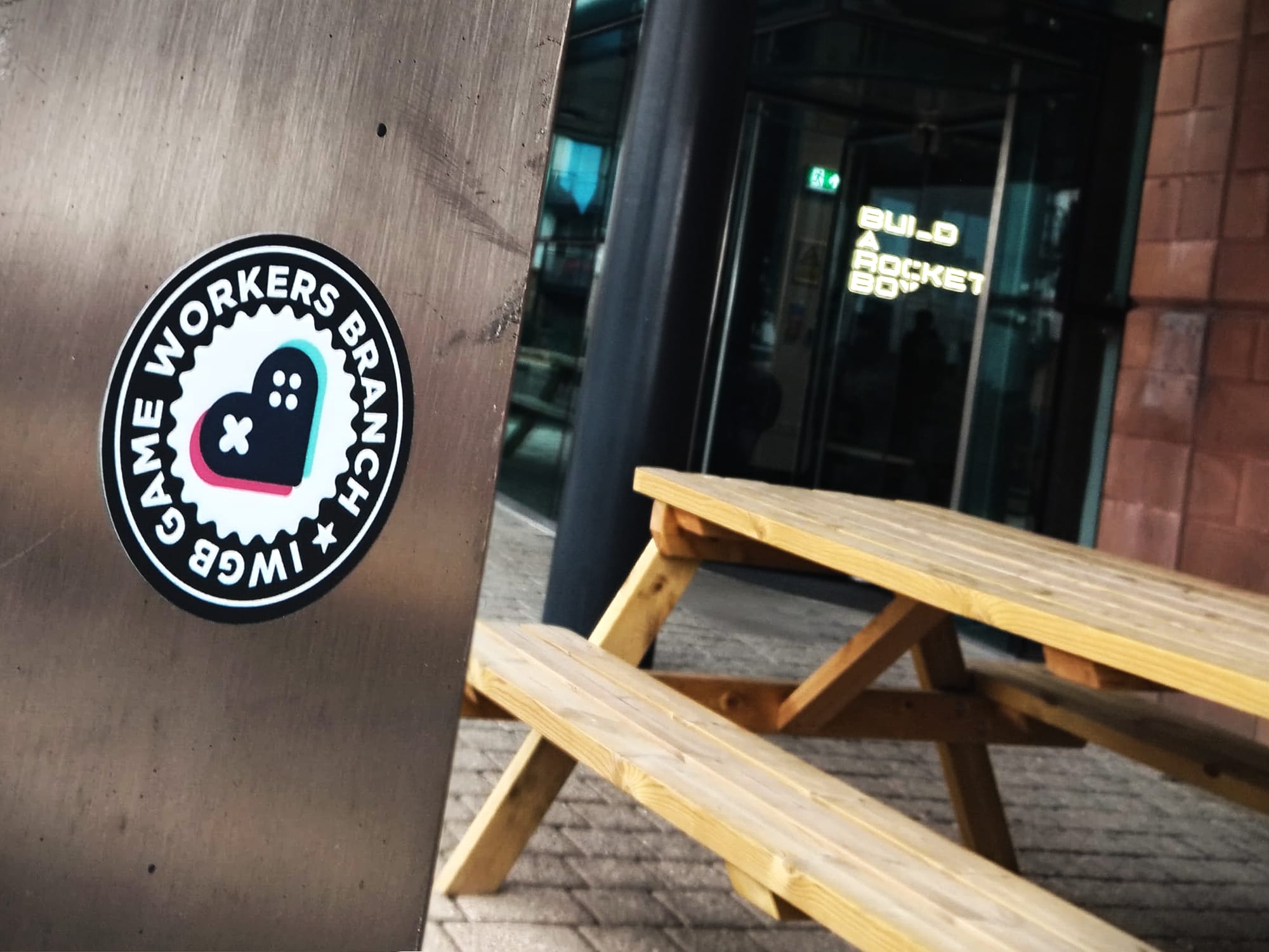 An IWGB Game Workers sticker appears in front of the Build a Rocket Boy office. A neon sign depicting the name of the studio appears in the background.